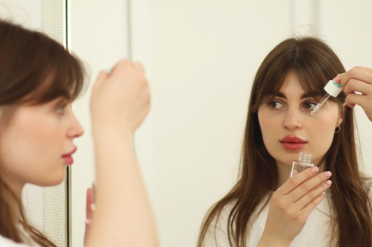 Services A young woman applying skincare serum in front of a mirror indoors. Perfect for beauty and self-care themes.